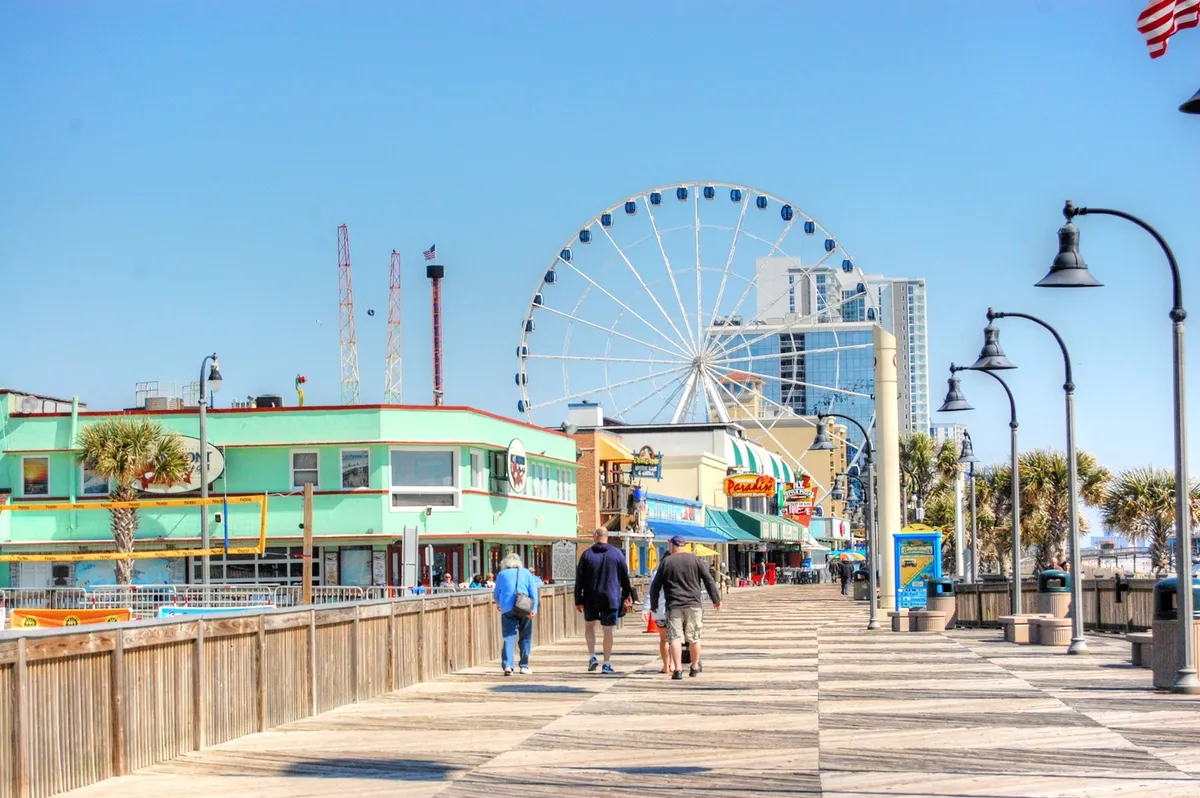 A vibrant exterior shot showing the Myrtle Beach Boardwalk with its colorful storefronts, palm trees, a Ferris wheel, and people strolling, shopping, and enjoying the festive atmosphere. The photo captures energy and opportunity for group outings.