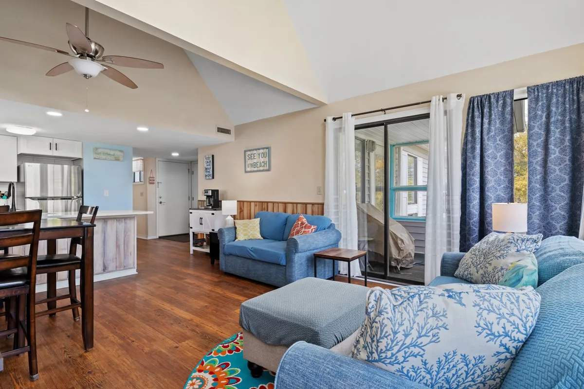 This image features a bright, open-concept living area with blue couches, a wooden dining table, decorative pillows, and a sliding glass door leading to an outdoor space. Sunlight streams in, creating a lively, inviting atmosphere ideal for relaxing or socializing.