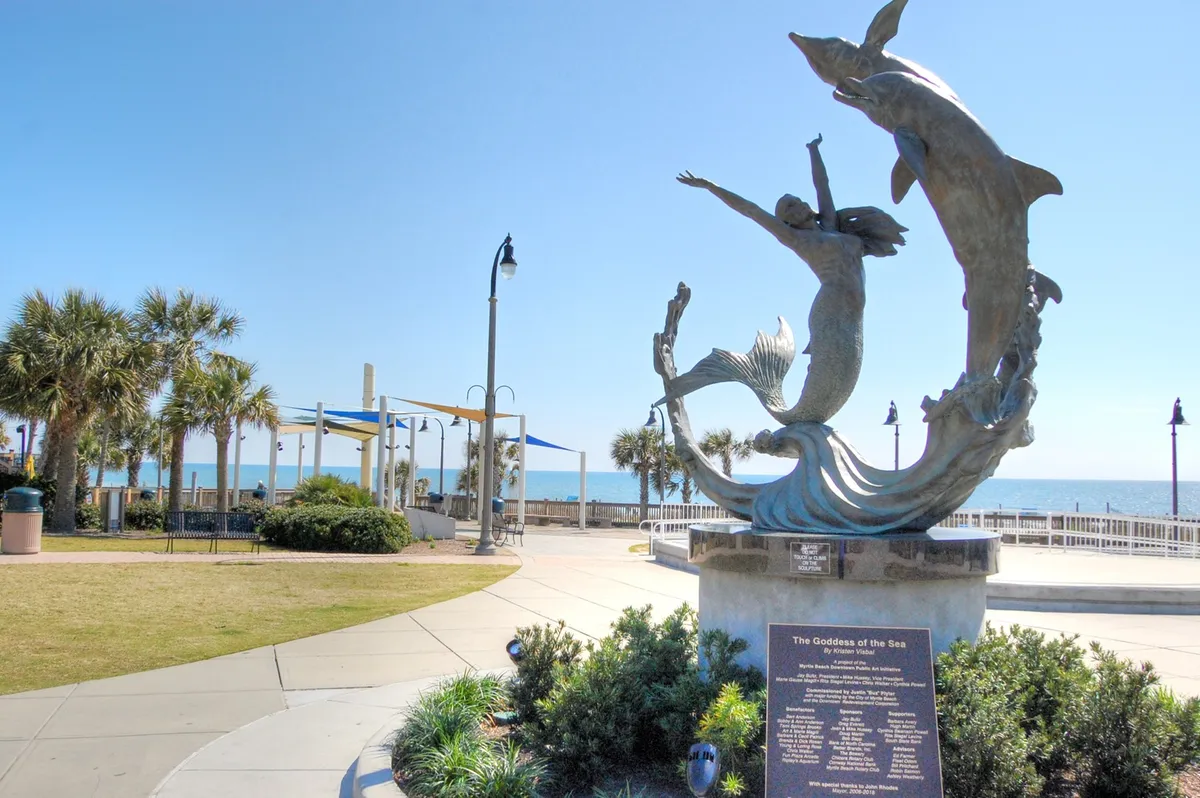 A scenic shot featuring the striking Goddess of the Sea sculpture, palm trees, the boardwalk area, and the ocean beyond. The image evokes a sense of place and adventure, inviting exploration and shared experiences along the coast.