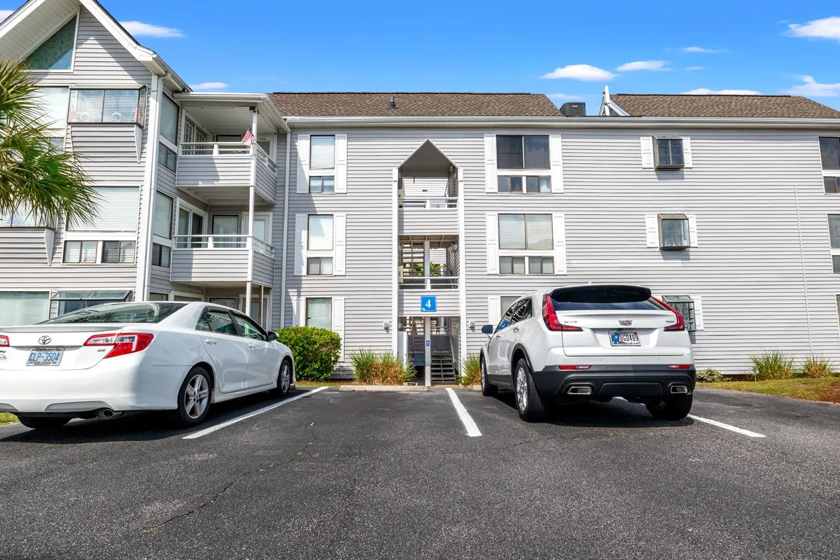 This exterior image captures the main entrance to the Arcadian Dunes building, with a 'number 4' sign clearly visible, two parked cars, and a welcoming atmosphere as people come and go from the apartments. The scene highlights the sense of community and accessibility at the property.
