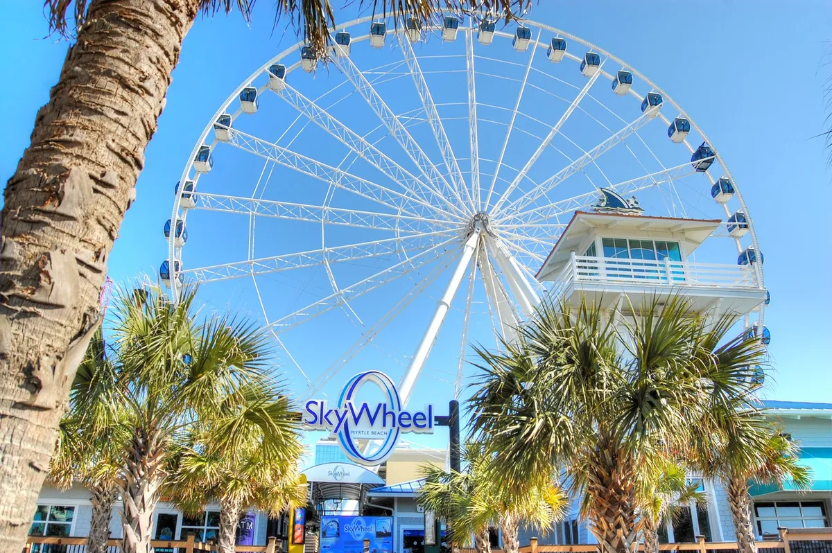 The SkyWheel stands tall against a bright sky, with palm trees and an inviting entrance sign in the foreground. This lively scene captures the excitement of a classic Myrtle Beach landmark, where visitors can enjoy panoramic views and create lasting memories.