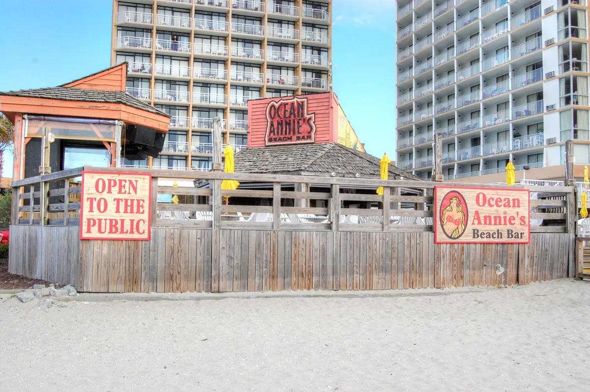 This exterior image features the iconic Ocean Annie's Beach Bar sign, a wooden beachfront structure, and a lively beach setting. The scene captures the beach bar's inviting vibe with people socializing, enjoying drinks, and the ocean as a backdrop.