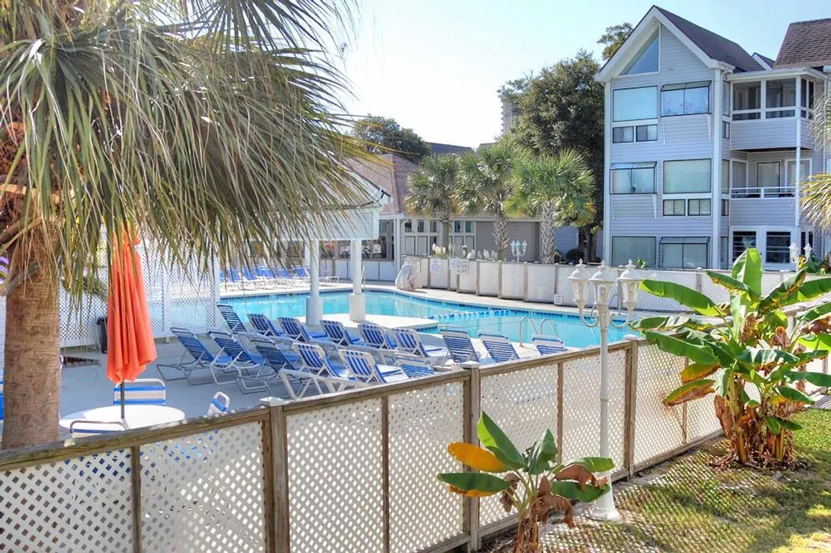 A vibrant outdoor resort pool area surrounded by palm trees and a building facade. Lounge chairs line the pool, inviting guests to sunbathe or read, while the space feels lively and inviting for both relaxation and socializing.