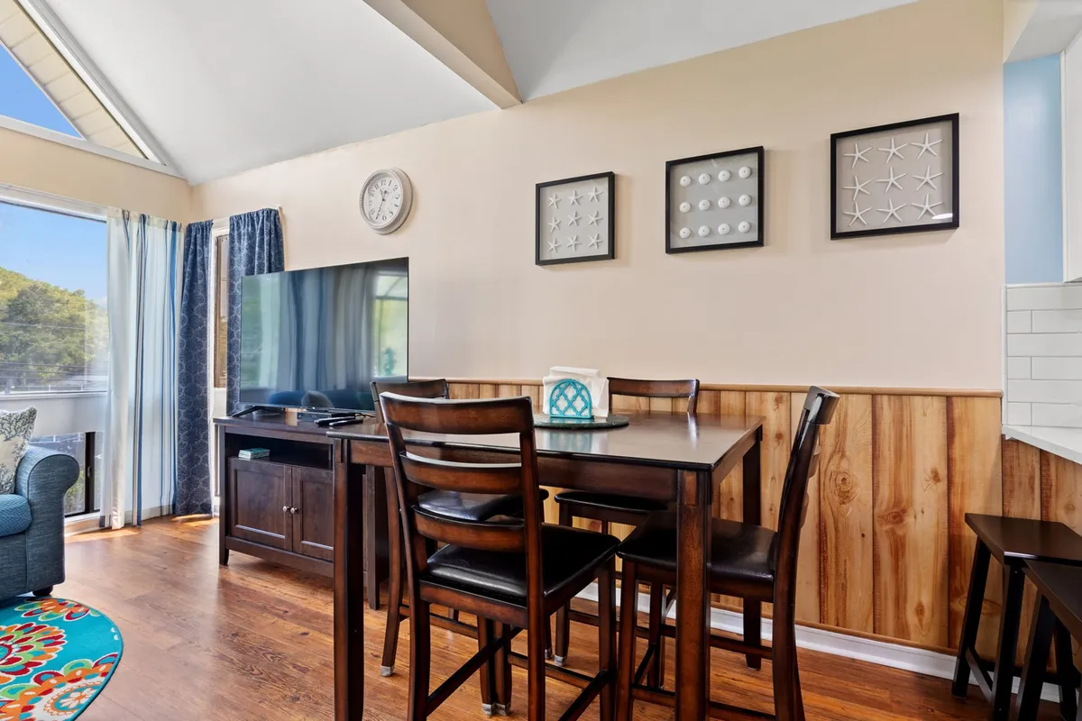 A cozy dining area with a table and chairs set for gathering, finished with a television, decorative wall art, curtains, and a rug. The setup invites group meals, celebrations, or casual conversations, fostering togetherness.
