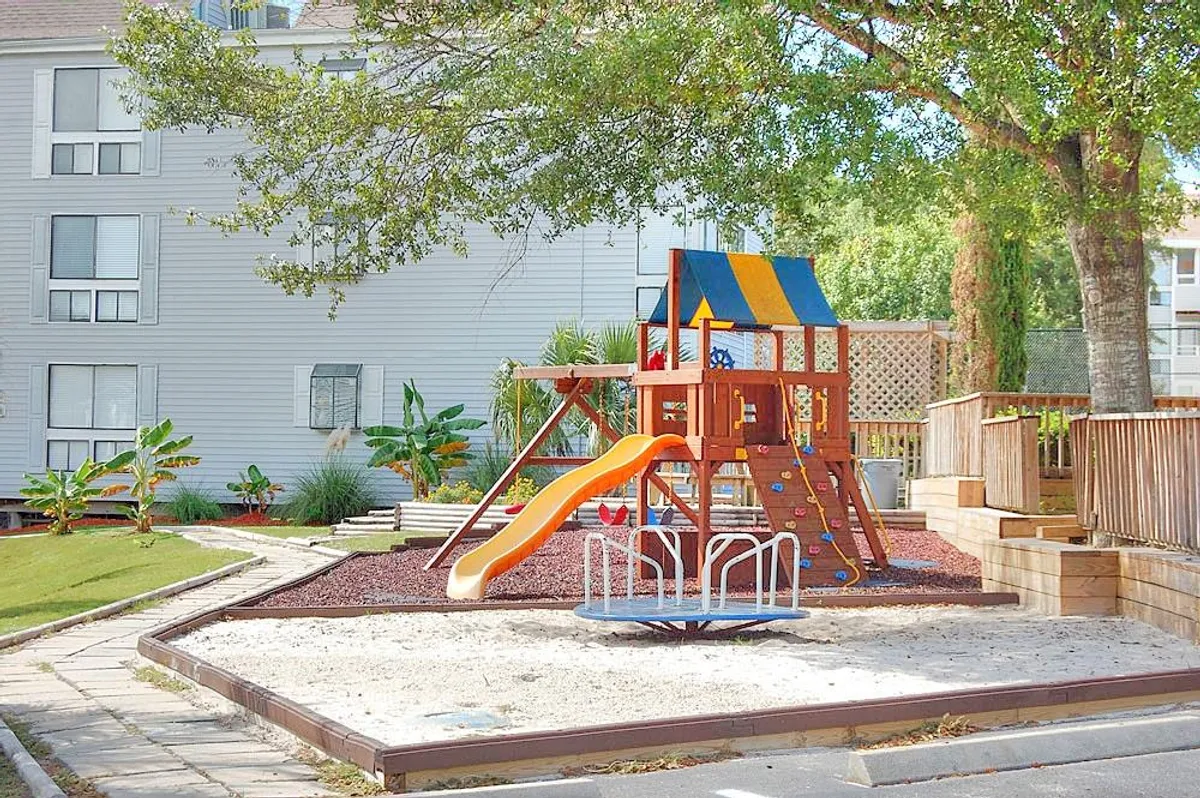 A vibrant playground area surrounded by green landscaping, featuring play equipment like a slide and merry-go-round. Children are seen playing, while parents and families gather nearby, highlighting the property’s appeal to families with young kids.
