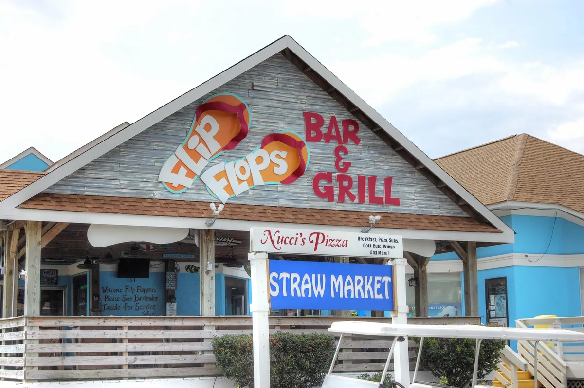 A festive exterior view of the 'Flip Flops Bar & Grill' sign, surrounded by vibrant banners and neighboring restaurant and shop signs. The lively street scene hints at on-site dining and socializing opportunities.