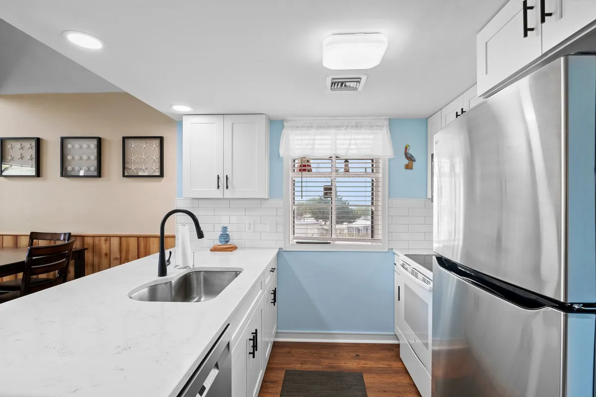 A bright, welcoming kitchen featuring a central island, vibrant blue accent wall, stainless steel appliances, and abundant natural light streaming in through a window. The island and spacious counters provide ample workspace, inviting multiple people to cook or gather together.