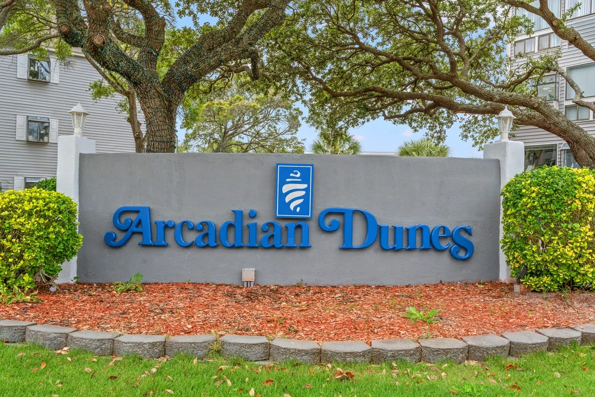 An exterior shot of the Arcadian Dunes sign framed by lush greenery and manicured landscaping, creating an inviting entry point for arriving guests. The scene sets a welcoming tone and hints at the condo’s central location.